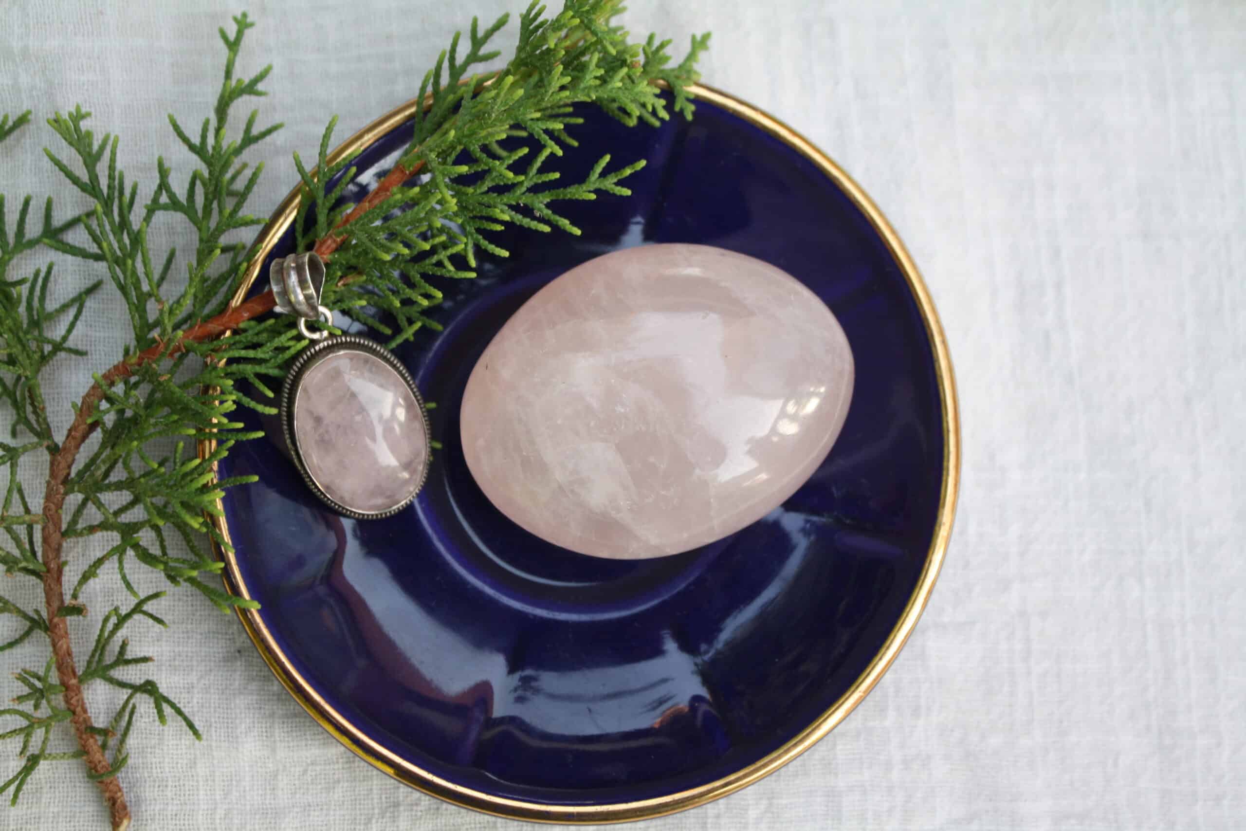 Polished rose quartz palmstone and necklace pendant on a dark blue dish with a twig
